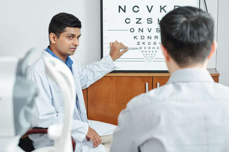 Indian optometrist pointing at whiteboard with letters and checking the eyesight of his patient at hospitalの写真素材