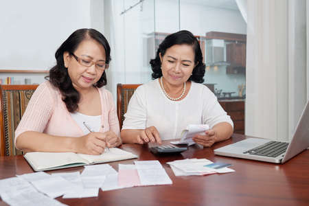 Two Asian mature women sitting at the table and working with home finances they checking their accounts on calculator and making notes in notepadの写真素材