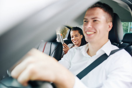 African woman smiling while sitting on back seat and using tablet pc with young driver driving his...