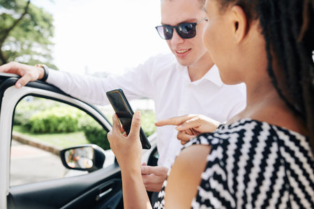 Young African woman showing online order on her mobile phone to a driver while they standing near...