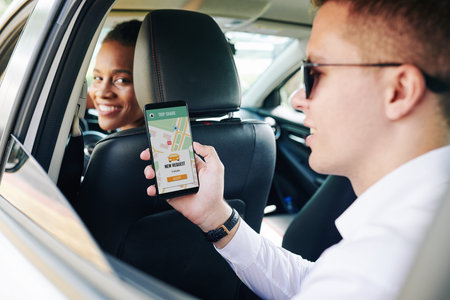 Young man wearing sunglasses holding mobile phone with car sharing app while sitting on back seat...