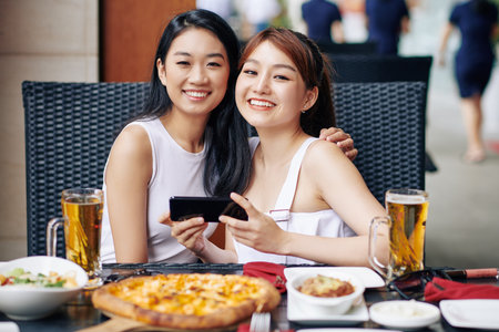 Portrait of two Asian women embracing and smiling at camera while using mobile phone during their...