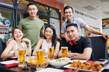 Portrait of Asian young friends smiling at camera while sitting at the table with glasses of beer...