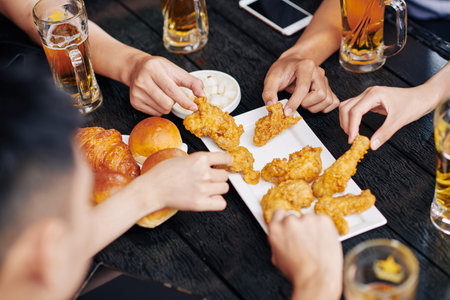 Close-up of people sitting at the wooden table drinking beer and eating fried chicken together in...