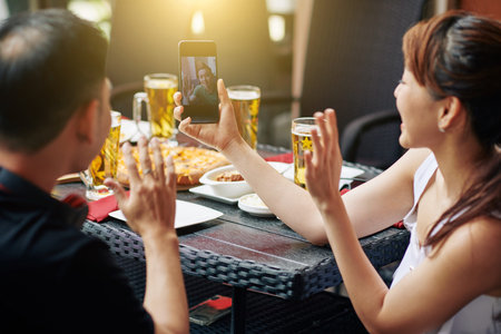 Rear view of young Asian couple holding mobile phone and have online conversation with their friend...
