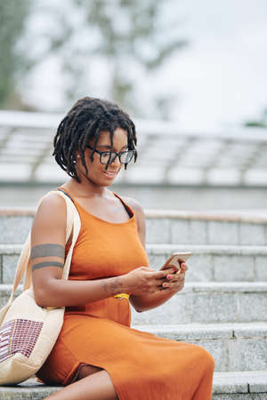 African happy woman in eyeglasses reading a message on mobile phone while resting on stairs outdoorsの写真素材