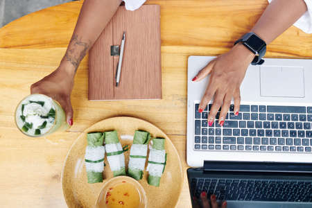 Close-up of woman sitting at wooden table have lunch and typing on keyboard of laptop computerの写真素材