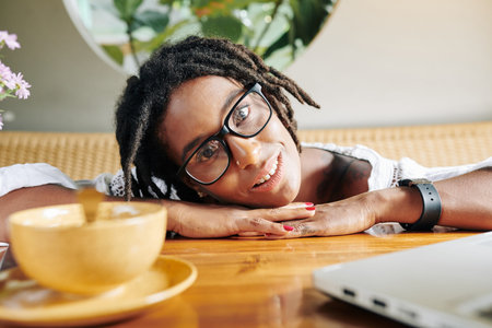 Portrait of African young woman in eyeglasses lying on the wooden table and smiling at camera in cafeの写真素材