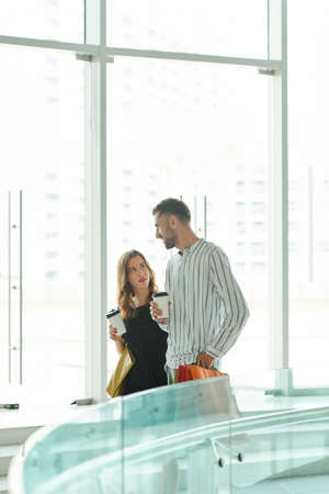 Serious pretty young woman with take away coffee looking at her talking boyfriend when they are standing on escalator in shopping mallの写真素材