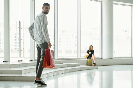 Attractive young woman resting after making purchases, her boyfriend with shopping bag walking to...