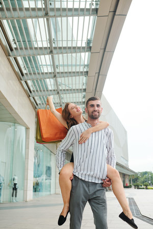 Cherful handsome young man giving piggyback ride to girlfriend after shopping in mall togeter