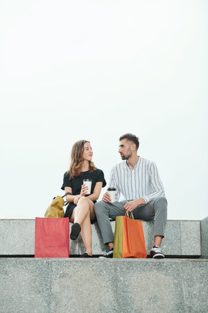 Positive young man and woman sitting on granite steps, drinking coffee and discussing sales and...