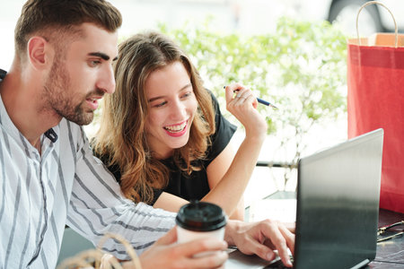 Cheerful boyfriend and girlfriend sitting at outdoor cafe with laptop and hot drinks and making...