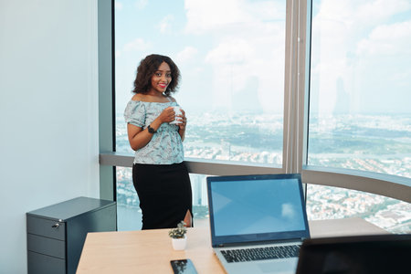 Shy smiling young Black businesswoman drinking coffee during short break at her fist day at work