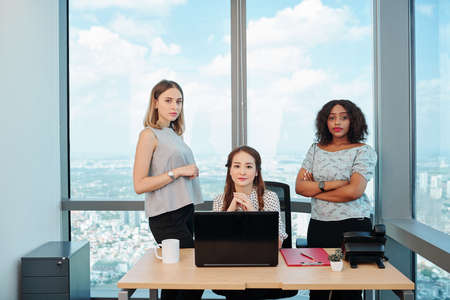 Young Vietnamese businesswoman sitting at office desk with opened laptop, her confident coworkers standing aroundの写真素材