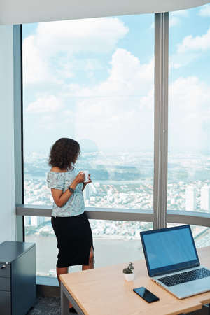 Pensive young Black businesswoman drinking cup of coffee and looking at big city through panoramic office windowの写真素材