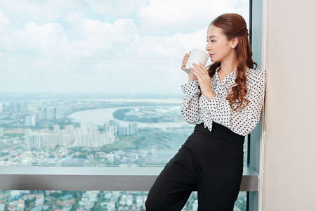 Pensive young pretty entrepreneur standing at big office window, enjoying cup coffee and city viewの写真素材