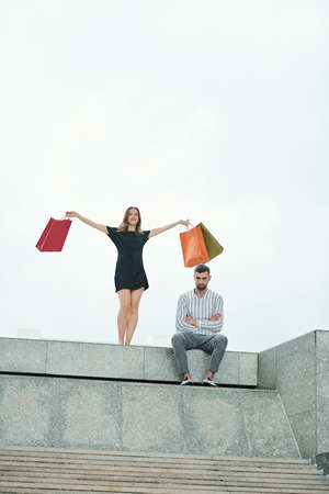 Unhappy man sitting outdoors with arms folded when his excited young girlfriend with many shopping-bags standing behindの写真素材