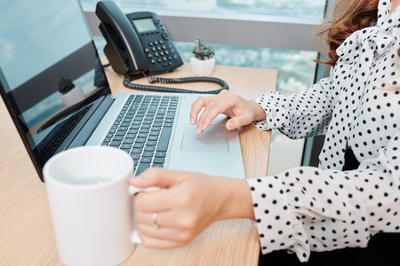 Hands of businesswoman drinking big mug of coffee and working on laptop in office