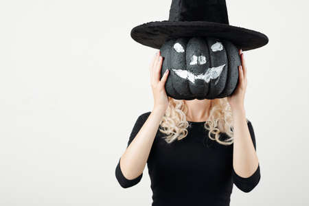 Young woman holding black pumpkin painted like jack-o-lantern in witch hat in front of her headの写真素材