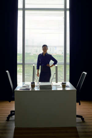 Young confident Asian businesswoman standing at office desk with hand on her waist and looking at cameraの写真素材