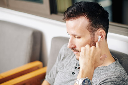 Serious bearded man using earbuds when watching show episode on laptop in cafe