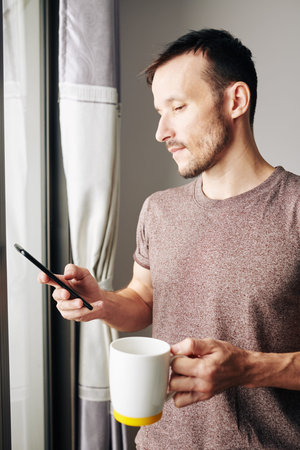 Smiling attractive young man in casual t-shirt holding big cup of tea and reading text messages from...
