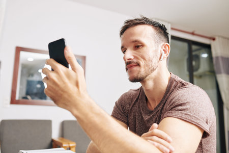 Young man wearing wireless earbuds when watching music video on smartphone screen