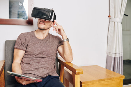 Man with tablet computer in hands using virtual reality headset when sitting on chair at home
