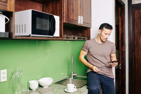 Man sitting on counter at kitchen sink and ordering food delivery via mobile applicationの写真素材