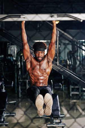 Shirtless determined fit young Black man doing hanging leg raise to strenghthen abs and core