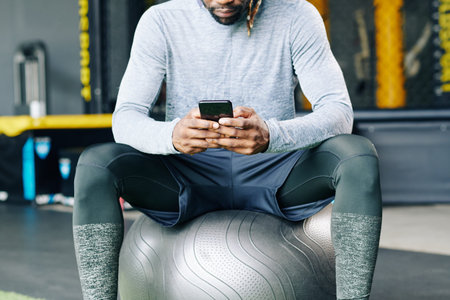 Cropped image of young sportsman sitting on big fitness ball and checking health application on...