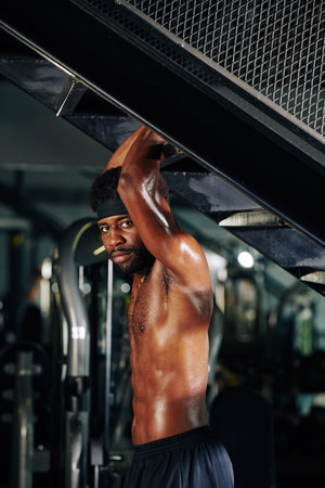 Side view of serious sweaty fit Black man standing at staircase in gym and looking at camera