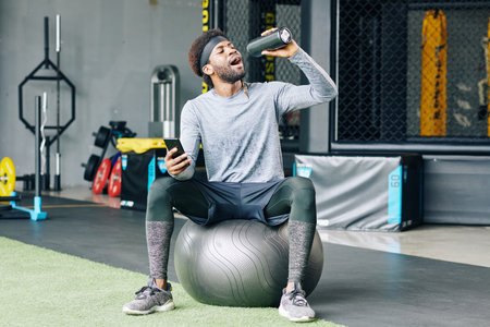 Sportsman with smartphone in hand resting on fitness ball between sets and drinking fresh water