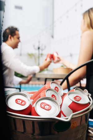Cans of soft drinks in ice bucket, cheerful partying people in background