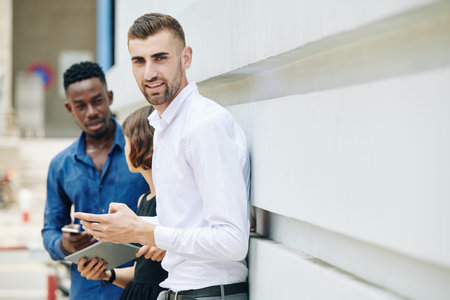 Portrait of handsome positive entrepreneur in white shirt standing outdoors with his colleagues and smiling at cameraの写真素材