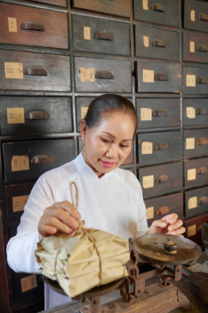 Positive traditional Asian apothecary worker putting plummet on scales and weighting packages with...