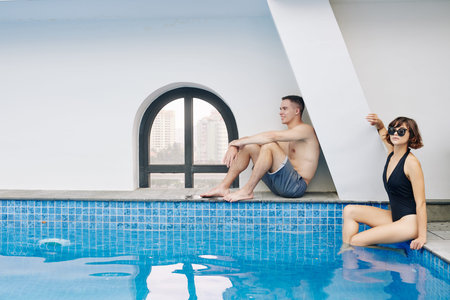 Young beautiful man and woman sitting at swimming pool with blue transparent water