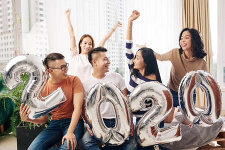 Excited friends with foil 2020 balloon having small New Year party at homeの写真素材