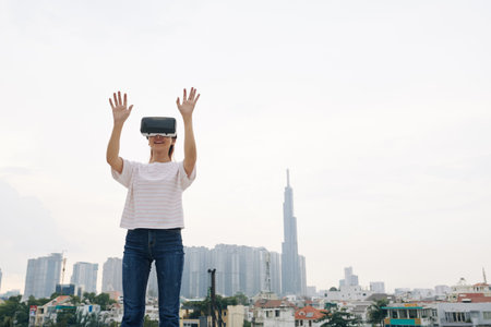 Happy young woman standing outdoors and raising hands to touch something when experiencing virtual...