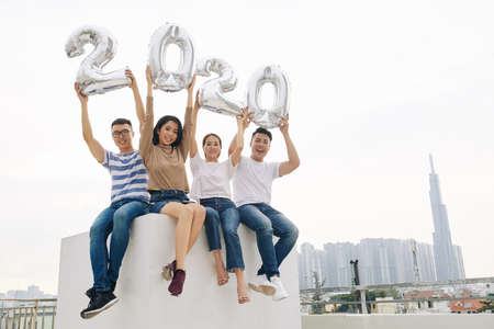 Joyful excited young Asian people sitting on rooftop and showing balloon figuresの写真素材
