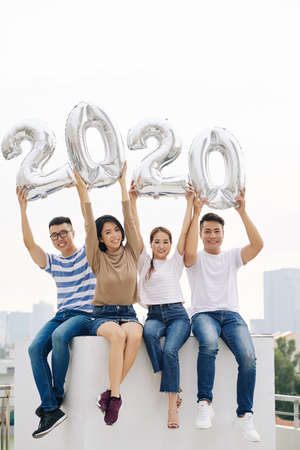 Group of positive young Vietnamese people showing 2020 foil balloons when sitting on the building roofの写真素材