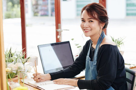Smiling pretty young florist taking notes in textbook after celling bouquet