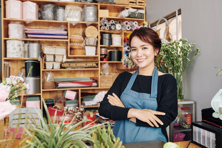Portrait of young positive florist in blue apron standing at counter with her arms folded and...