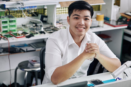 Portrait of young positive Asian repairman working at table at smartphone repair shop