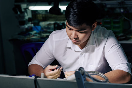 Pensive young Asian repairman looking at smartphone logic board in his hand
