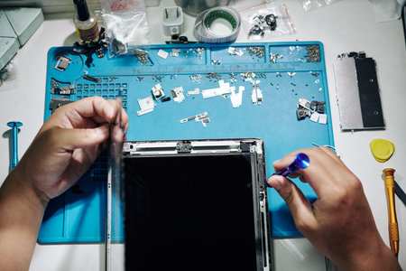 Repairman using tweezers and screwdriver when removing screen of tablet computer at his desk, view...