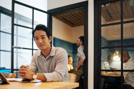 Portrait of handsome young successful entrepreneur in striped shirt sitting at office desk and smiling at cameraの写真素材