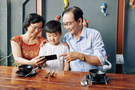 Senior Asian couple showing mobile application to grandson when they are sitting at cafe tableの写真素材