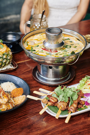 Thai spicy squid soup in metal pot and various seafood snacks on dinner table in cafeの写真素材